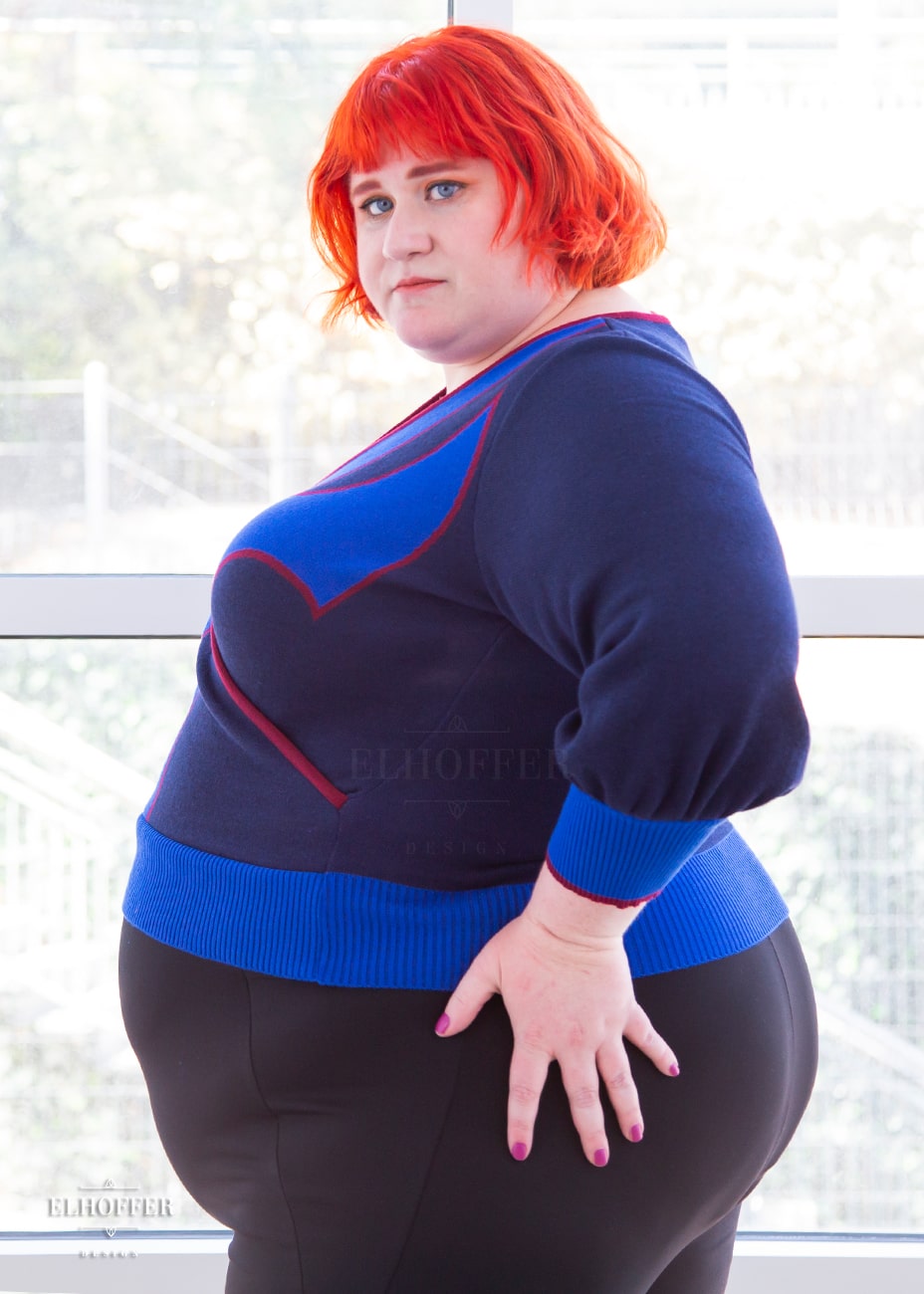 Logan, a fair skinned size 3XL model with bright orange hair and bangs, is wearing a crossover knit crop top. The base is a dark blue, details are bright blue with a red outline and form almost a t shape in the middle of the crop. The cuffs and bottom are the same bright blue of the detail and the crossover is piped in the same red.