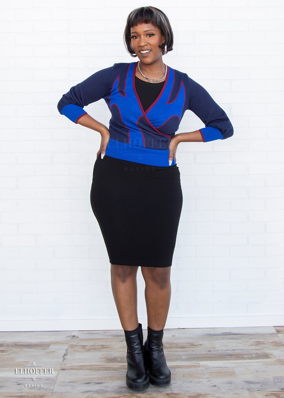 Lynsi, a medium dark skinned model with short black and white hair, is wearing a crossover knit crop top. The base is a dark blue, details are bright blue with a red outline and form almost a t shape in the middle of the crop. The cuffs and bottom are the same bright blue of the detail and the crossover is piped in the same red.