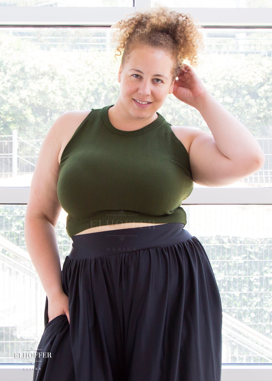 Anastasia (a medium fair skinned size extra large model with curly blonde hair) wears the olive green sleeveless high necked knit crop top. Its hem falls to her natural waist.