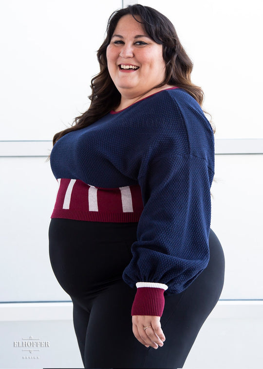 Alysia, a sun kissed skin size XL model with long brown hair, is wearing the red, white and blue knit oversized crop top with bishop sleeves. The sleeves and top of the crop are blue with a waffle knit, the waist is vertical red and white stripes. The cuffs are mainly red with a white detail. There is also a red detail at the neckline.