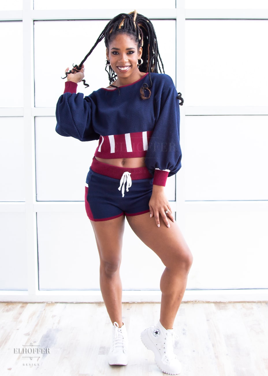 Krystina, a medium dark skinned S model with long braids in an updo, is smiling while wearing the red, white and blue knit oversized crop top with bishop sleeves. The sleeves and top of the crop are blue with a waffle knit, the waist is vertical red and white stripes. The cuffs are mainly red with a white detail. There is also a red detail at the neckline. She paired the sweater with a matching pair of blue and red knit shorts.
