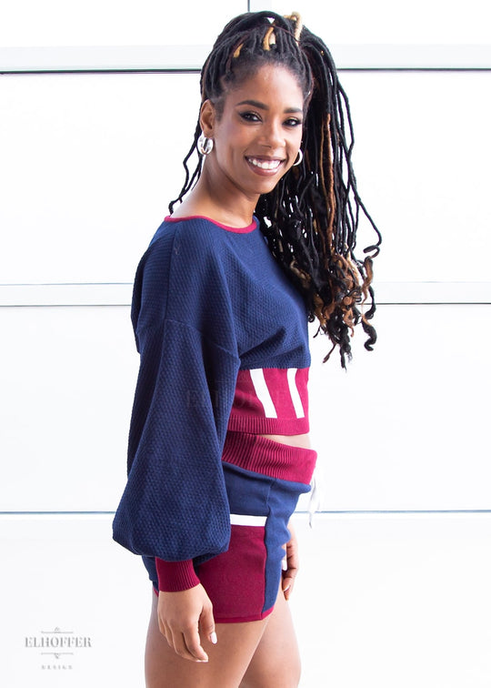 Krystina, a medium dark skinned S model with long braids in an updo, is smiling while wearing the red, white and blue knit oversized crop top with bishop sleeves. The sleeves and top of the crop are blue with a waffle knit, the waist is vertical red and white stripes. The cuffs are mainly red with a white detail. There is also a red detail at the neckline. She paired the sweater with a matching pair of blue and red knit shorts.