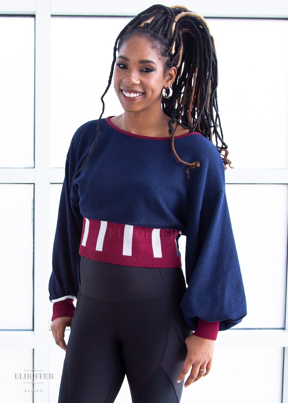 Krystina, a medium dark skinned S model with long braids in an updo, is smiling while wearing the red, white and blue knit oversized crop top with bishop sleeves. The sleeves and top of the crop are blue with a waffle knit, the waist is vertical red and white stripes. The cuffs are mainly red with a white detail. There is also a red detail at the neckline.