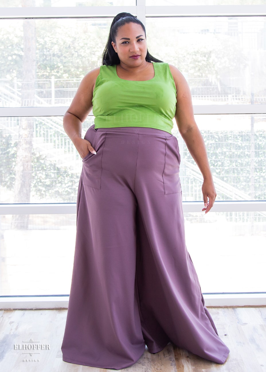 Tas, a medium skinned size 2XL model with long dark brown hair is wearing high waisted full palazzo pants with front pockets and an invisible zipper on the side seam in mauve.