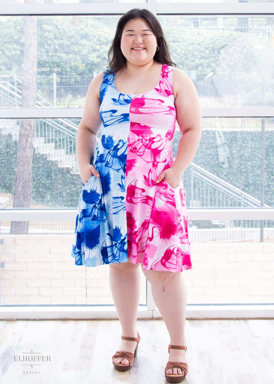 Ashley (a fair skinned size large / extra large model with long dark hair) wears the scoop neck sleeveless spandex knee length dress. The right half of the dress is a blue alcohol ink swirl pattern and the left half of the dress is a hot pink alcohol ink swirl pattern.