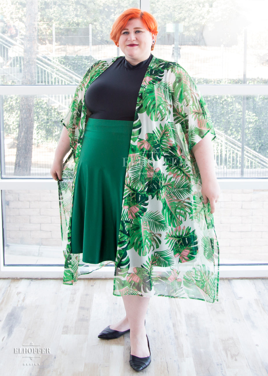 Logan, a fair skinned 3XL model with short bright orange hair, is wearing the open front sheer cover up. The cover up has a pattern of green tropical leaves with pink spots on a white background.