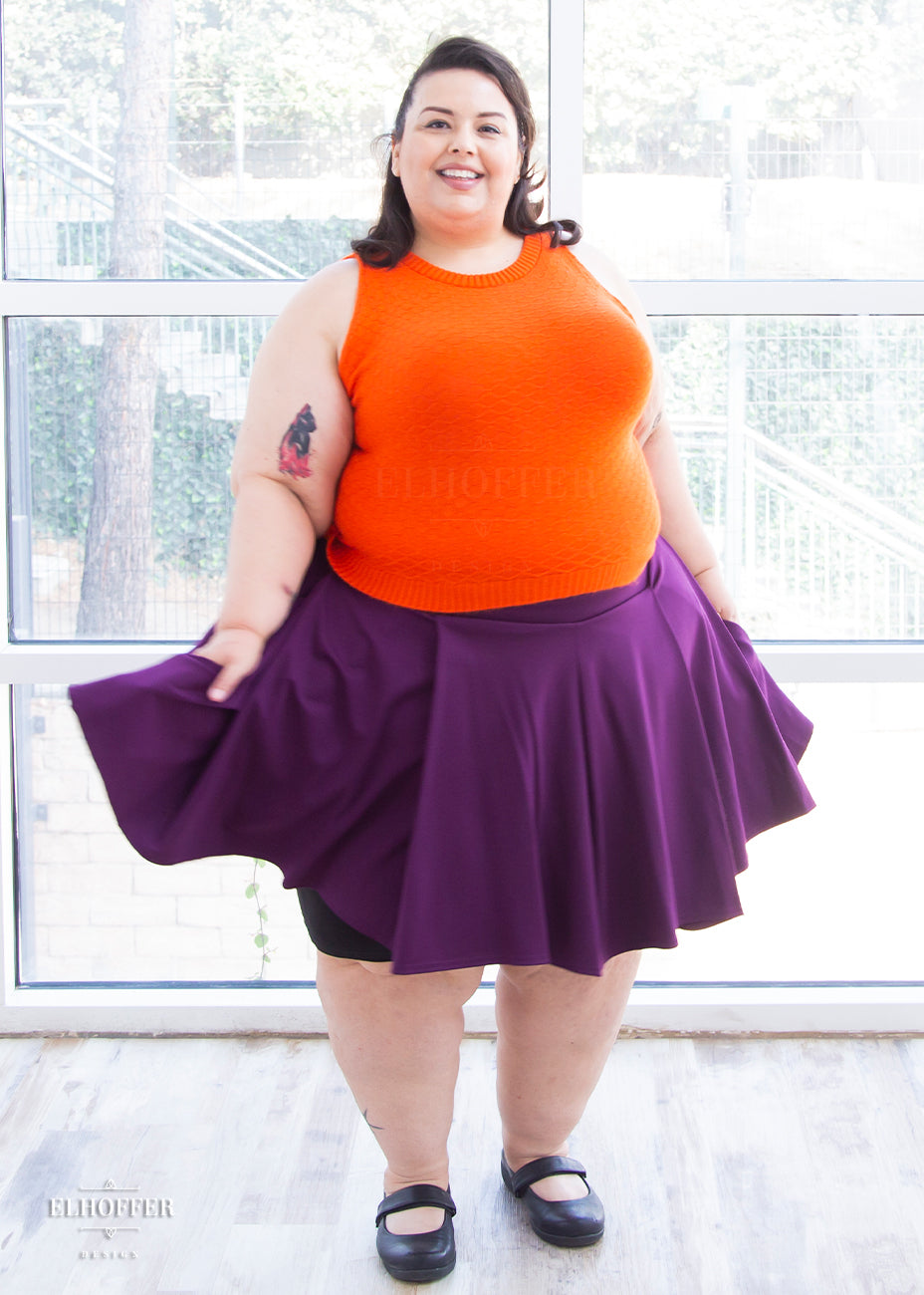 Angel (a fair skinned size 4XL model with brunette hair) models the knee length pleated plum purple skirt with pockets. She pairs it with a bright orange knit sleeveless Susan crop top.models the knee length pleated plum purple skirt with pockets. She pairs it with a bright orange knit sleeveless Susan crop top.