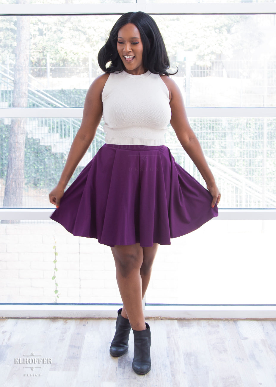 Lynsi (a dark skinned size M model with long dark hair) models the knee length pleated plum purple skirt with pockets. She pairs it with a stone beige knit sleeveless Susan crop top.