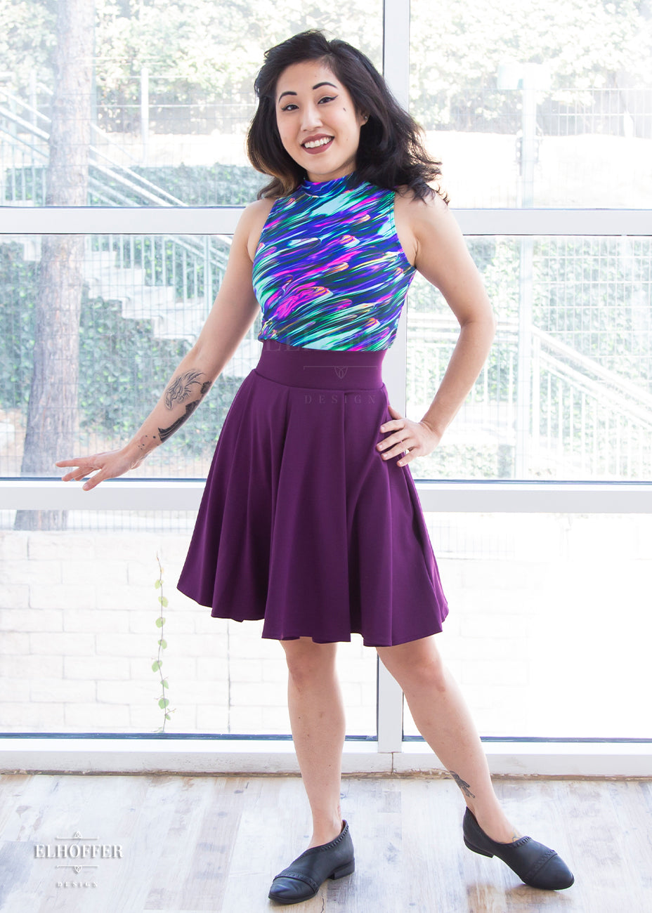 Kate (a medium fair skinned size extra small model with dark hair) models the knee length pleated plum purple skirt with pockets. She pairs it with a spandex sleeveless faux turtleneck Jes crop top in a colorful oil paint print. 