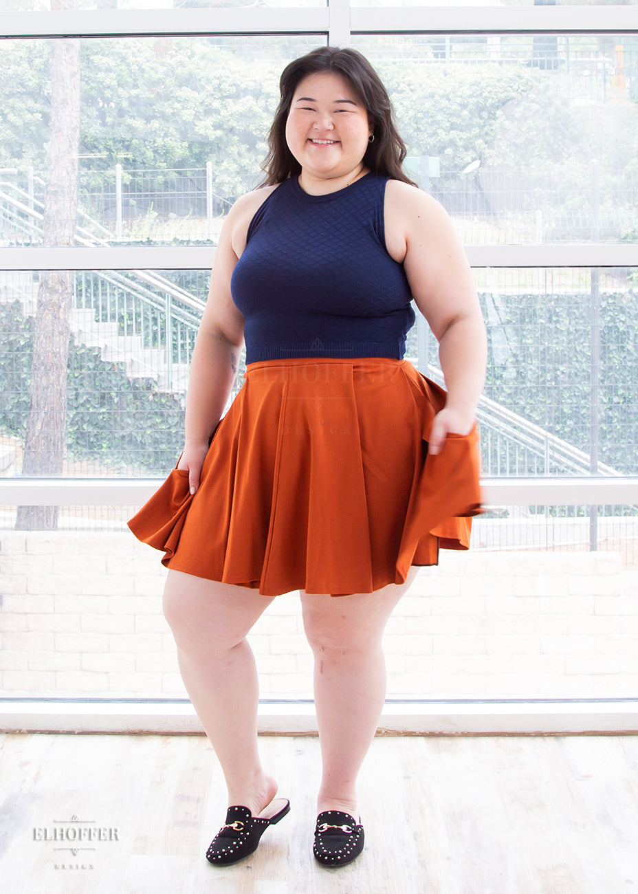 Ashley (a fair skinned size large / extra large model with long dark hair) wears the pleated skater length rust orange ponte skirt paired with a navy blue high neck sleeveless crop top.