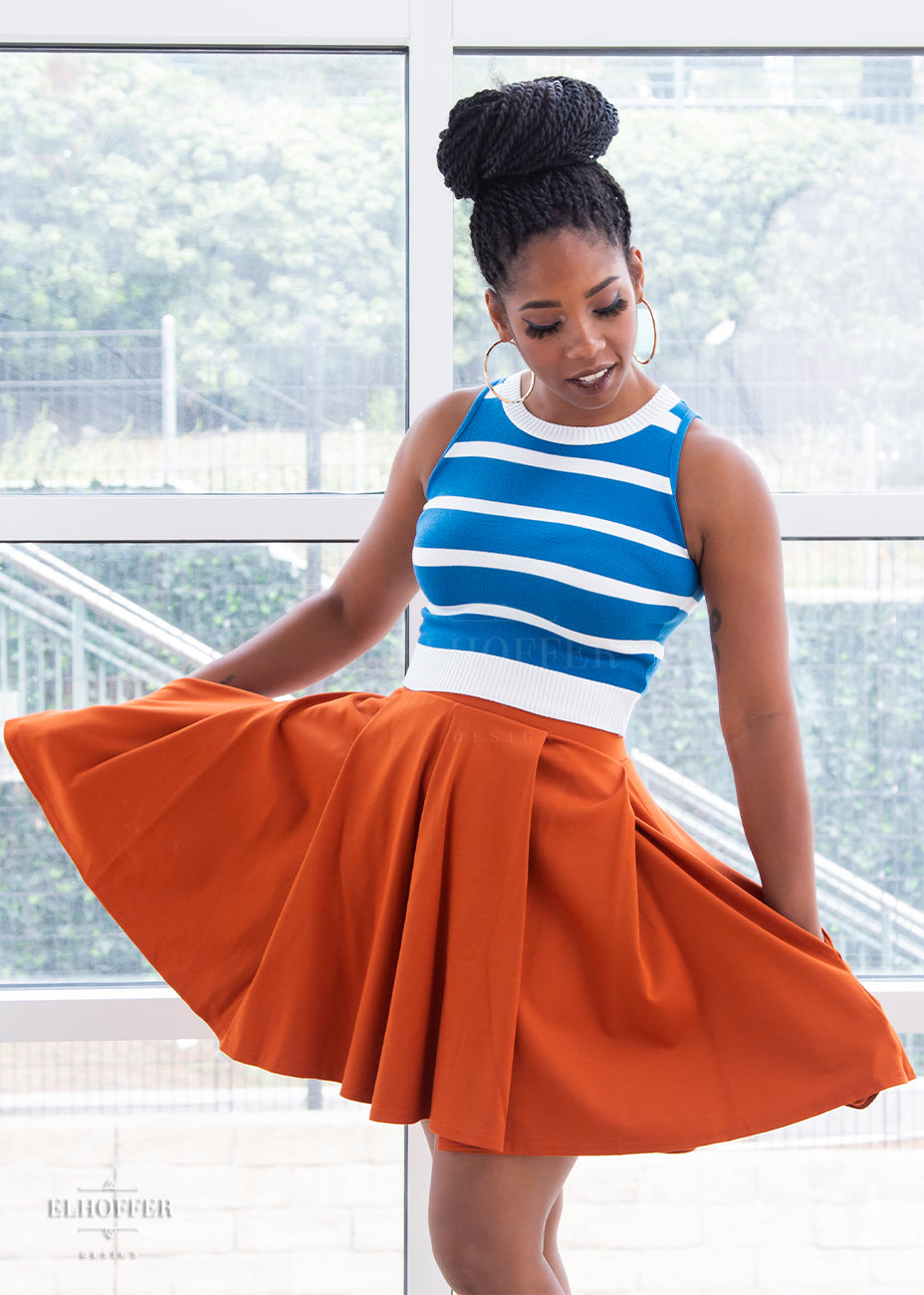 Krystina (a dark skinned size small model with long dark twist braids) wears the pleated skater length rust orange ponte skirt with her hands in the skirt's pockets.