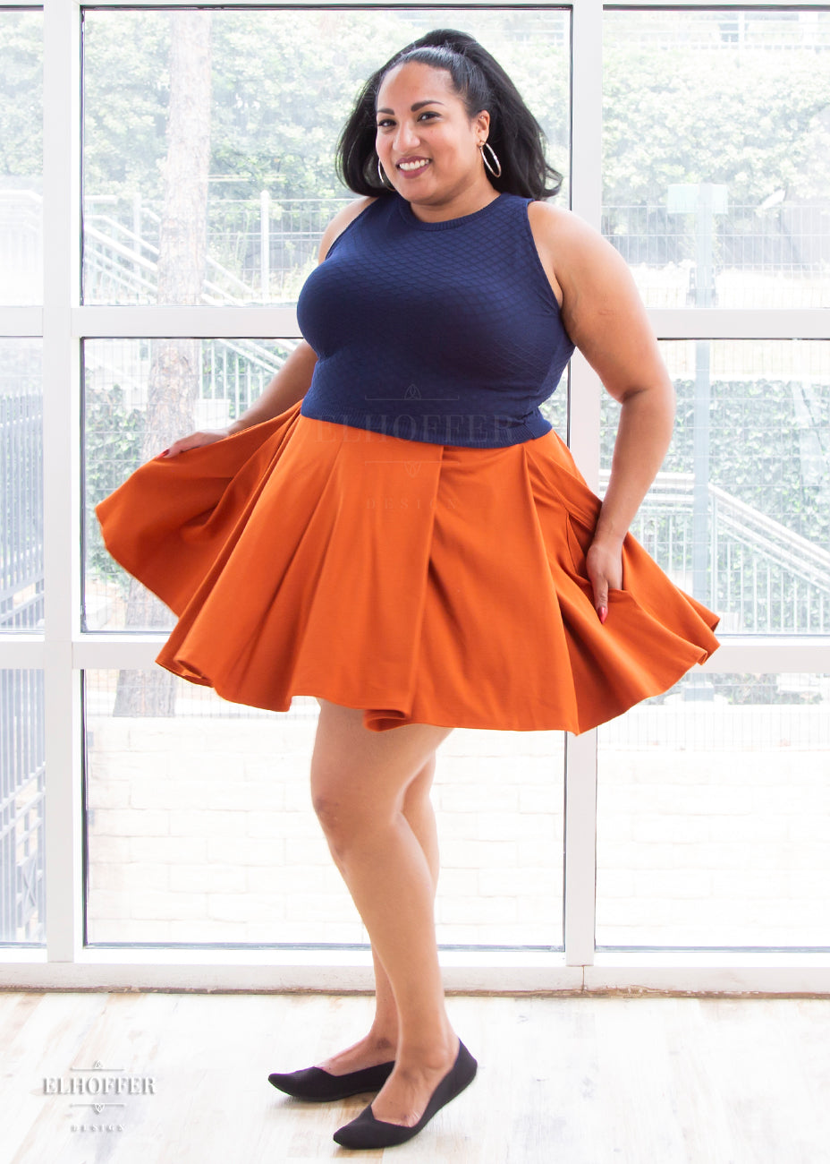 Tas (a medium skinned size 2X model with long dark hair in a ponytail) wears the pleated skater length rust orange ponte skirt with her hands in the skirt's pockets.