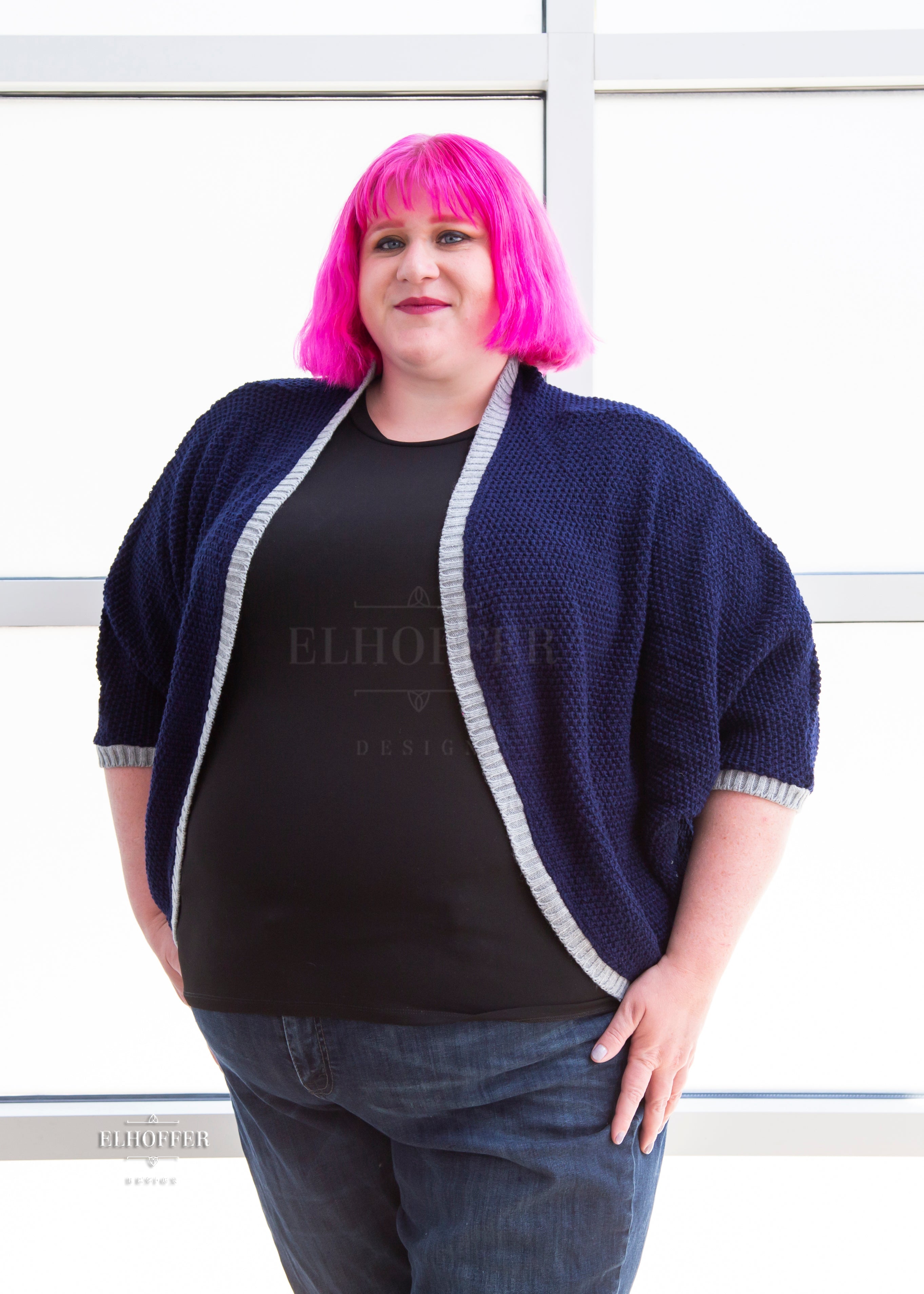 Logan, a fair skinned 3xl model with short bright pink hair with bangs, is wearing a dark blue loose knit dolman with light grey ribbing around edges and cuffs. The dolman features 3/4 length sleeves and a magical script design (circle with T crossing through the circle) on the back.