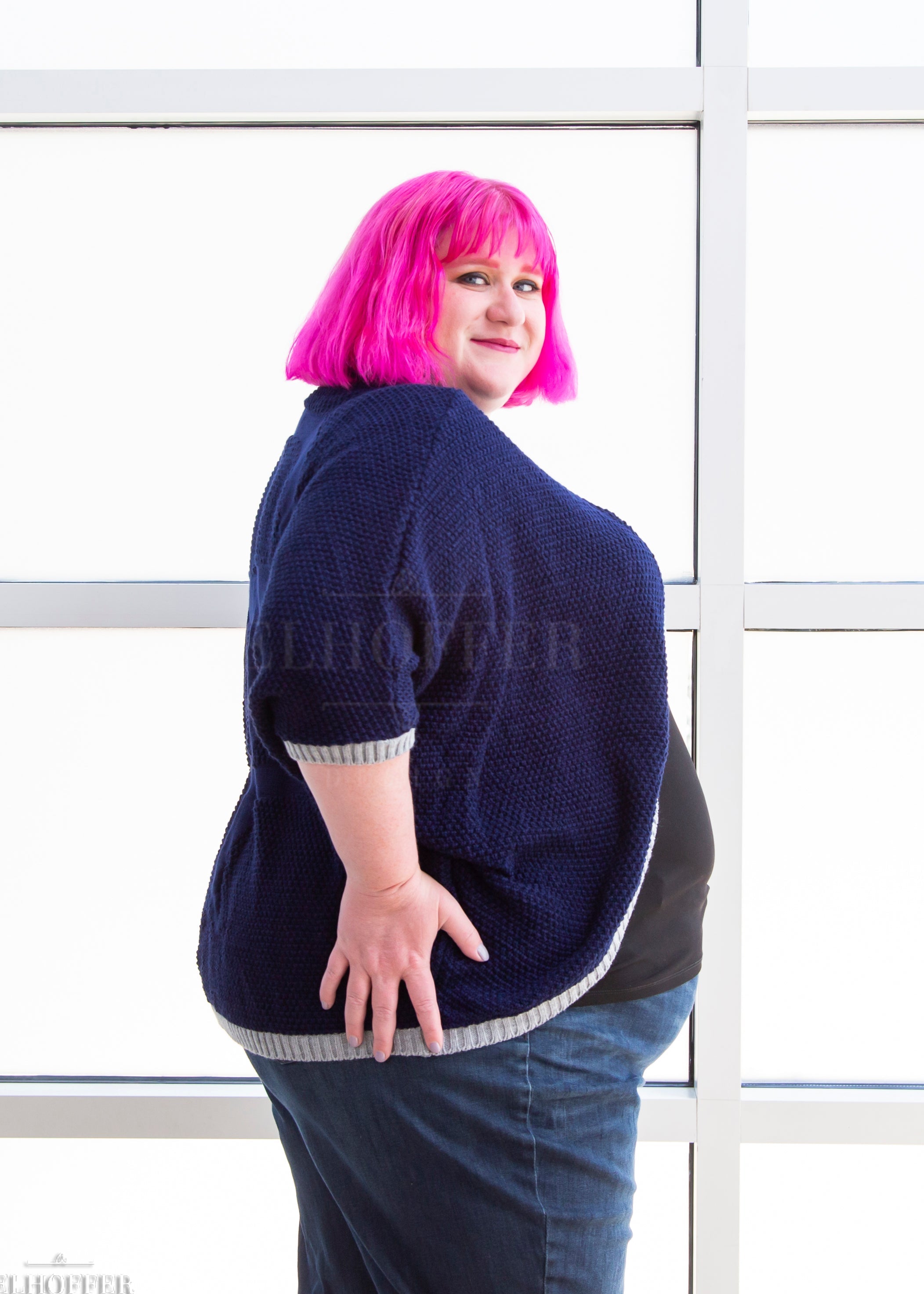 Logan, a fair skinned 3xl model with short bright pink hair with bangs, is wearing a dark blue loose knit dolman with light grey ribbing around edges and cuffs. The dolman features 3/4 length sleeves and a magical script design (circle with T crossing through the circle) on the back.