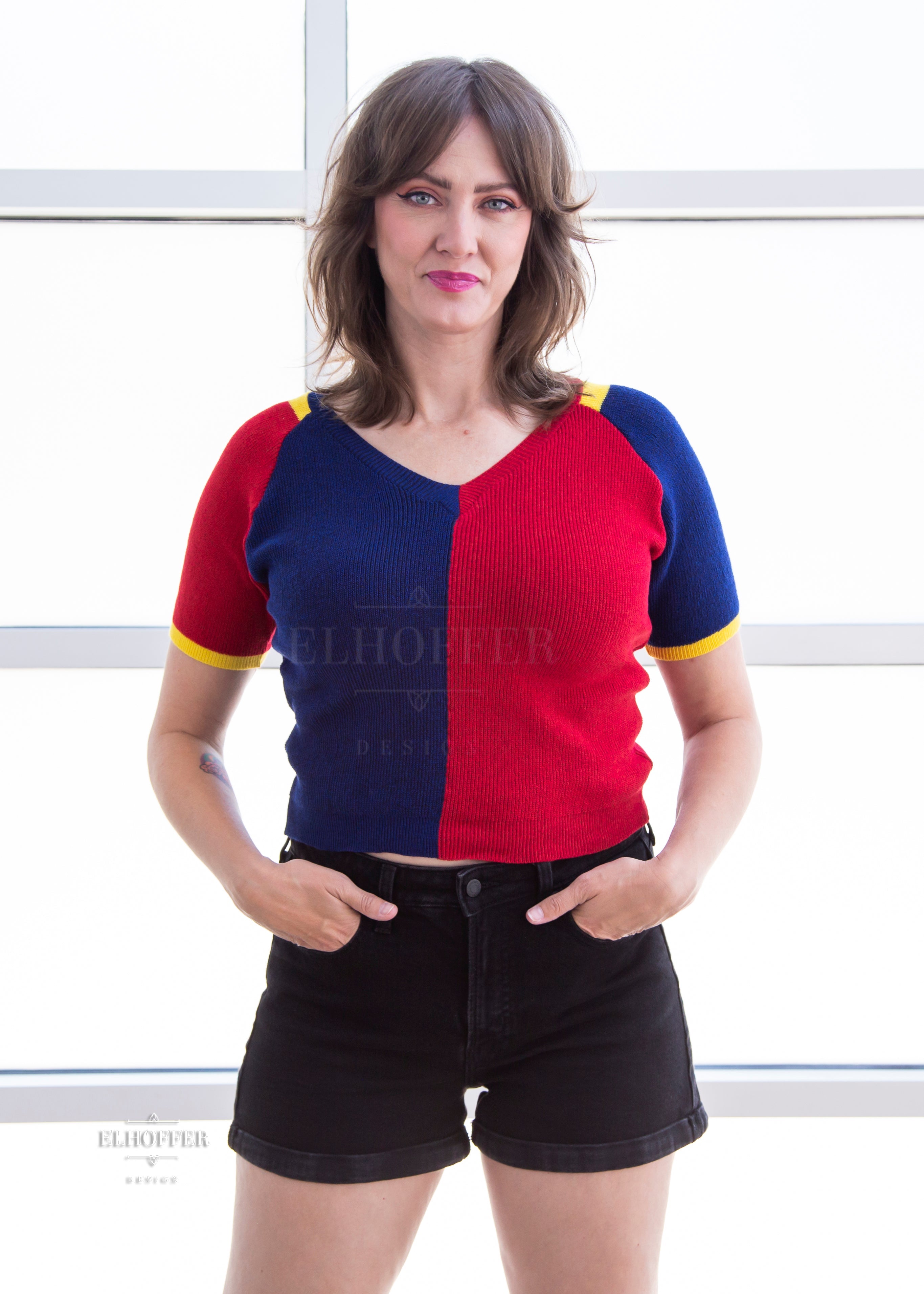 Ashley, a light olive skinned M model with shoulder length brown feathered hair, is wearing a short sleeve knit top with alternating blue and red colors. There is yellow detailing along the top of the shoulder and around the cuff of the sleeve.