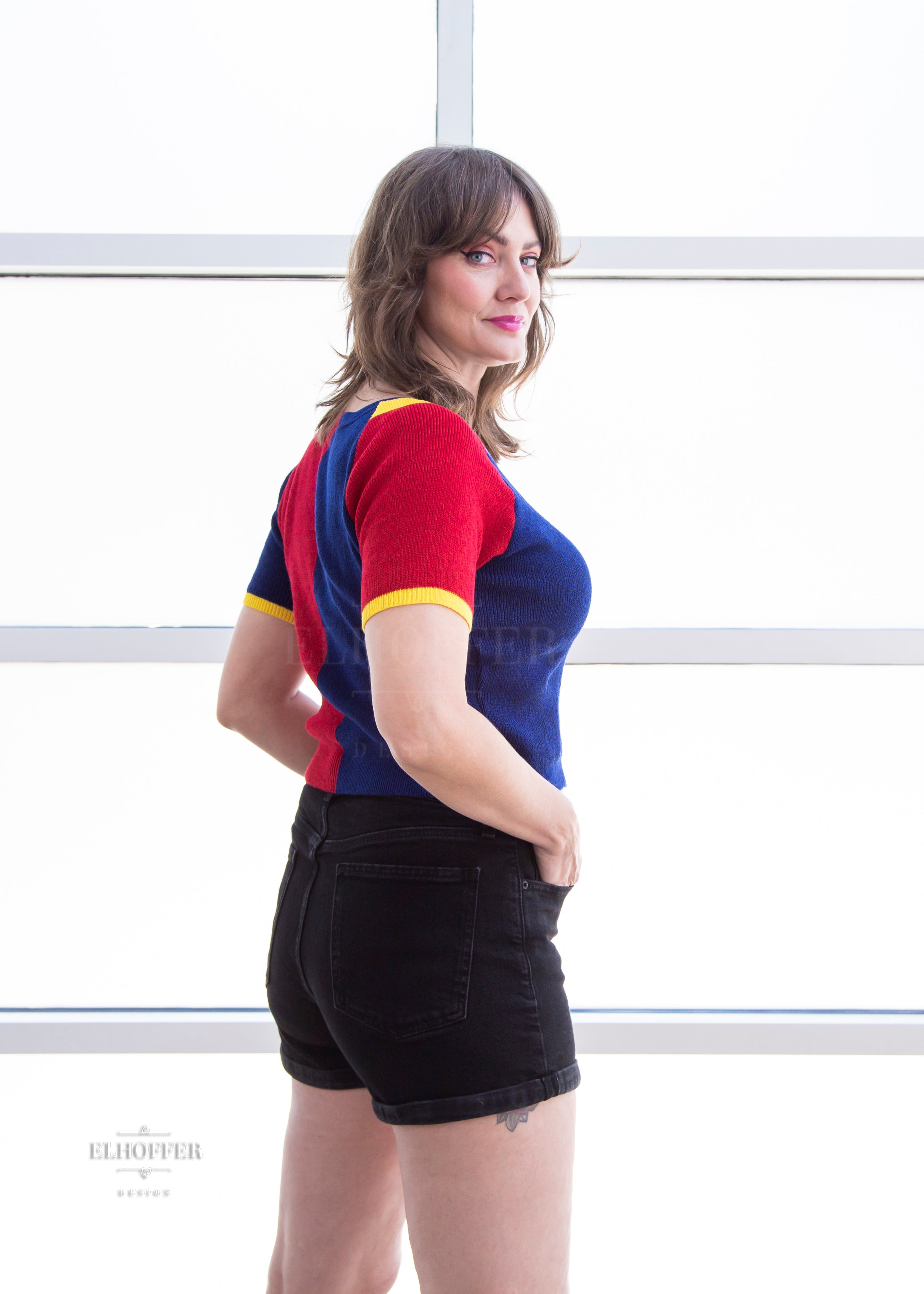 A side view of Ashley, a light olive skinned M model with shoulder length brown feathered hair, wearing a short sleeve knit top with alternating blue and red colors. There is yellow detailing along the top of the shoulder and around the cuff of the sleeve.