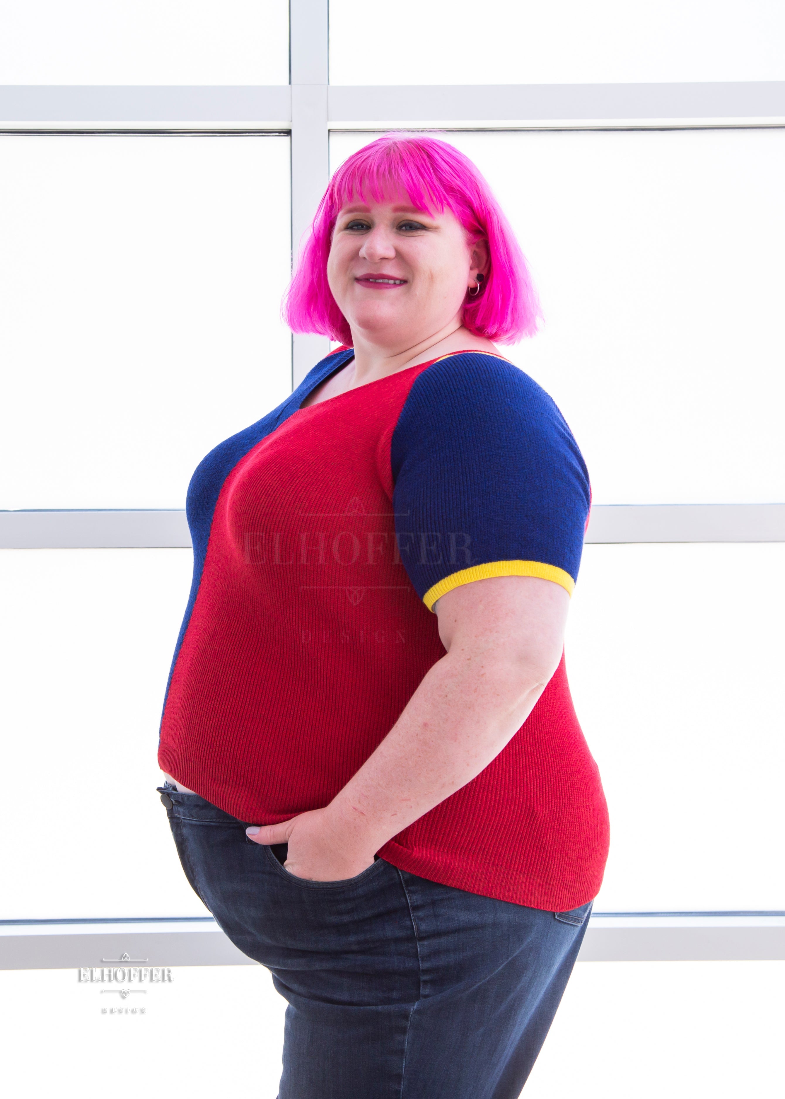 A side view of Logan, a fair skinned 3xl model with short pink hair with bangs, wearing a short sleeve knit top with alternating blue and red colors. There is yellow detailing along the top of the shoulder and around the cuff of the sleeve.