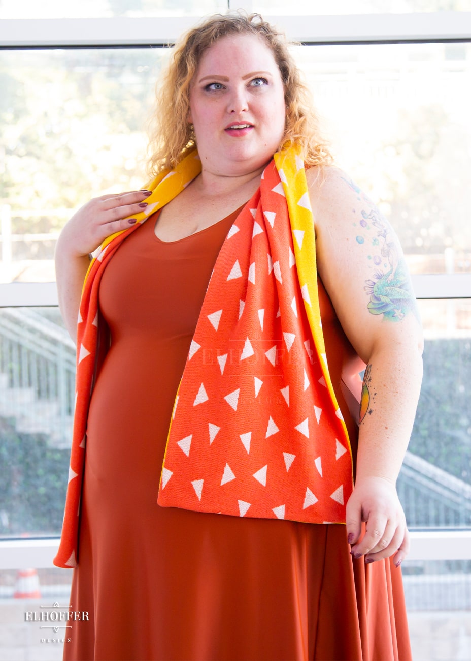 Bee, a fair skinned 3xl model with medium length curly blonde hair, is wearing a reversible knit scarf.  One side of the scarf is orange with white triangles and the other side is yellow with white triangles.