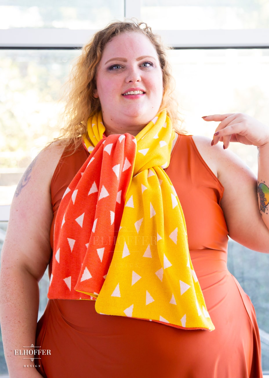 Bee, a fair skinned 3xl model with medium length curly blonde hair, is smiling while wearing a reversible knit scarf.  One side of the scarf is orange with white triangles and the other side is yellow with white triangles.