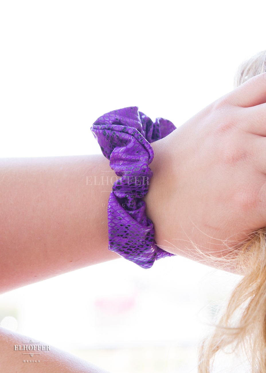 A shiny purple snake skin patterned scrunchie on a fair freckled wrist.