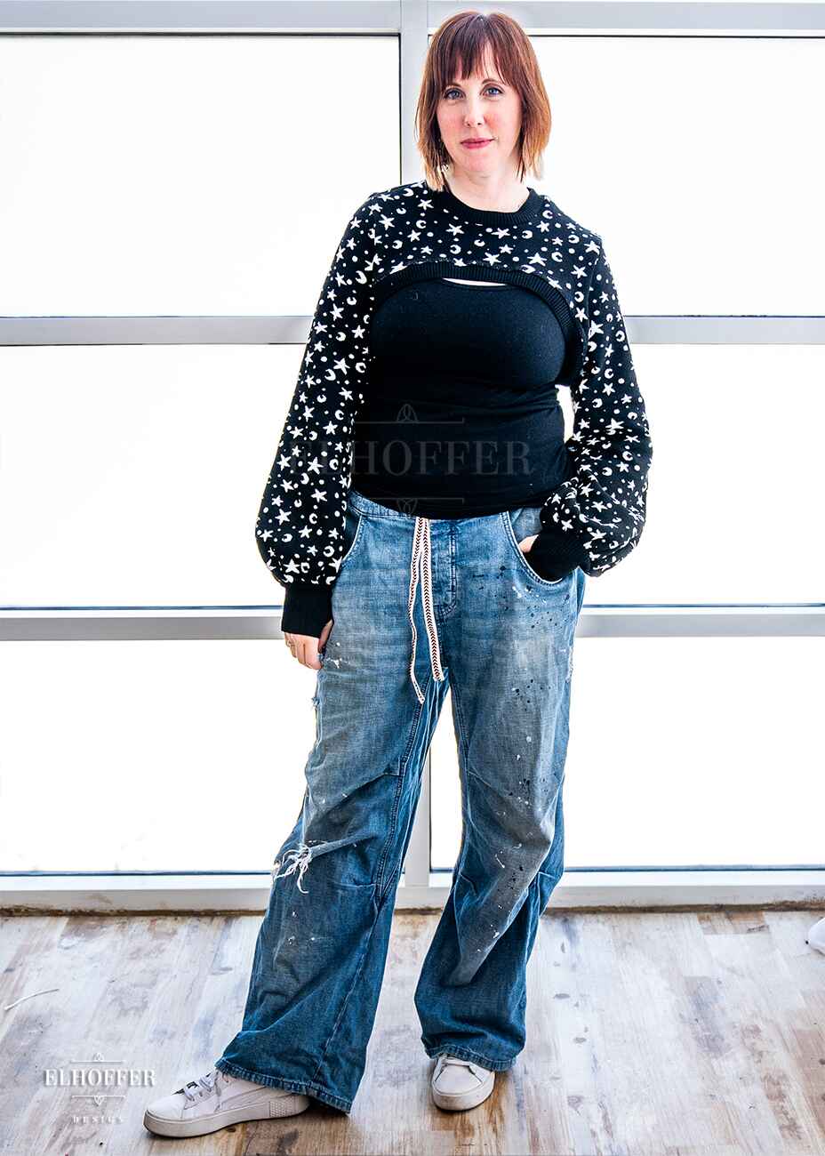 Jes, a fair skinned S model with short red hair and bangs, is wearing the L sample of a black crew neck super cropped knit shrug sweater with a white star and moon pattern, long billowing sleeves and thumbholes.
