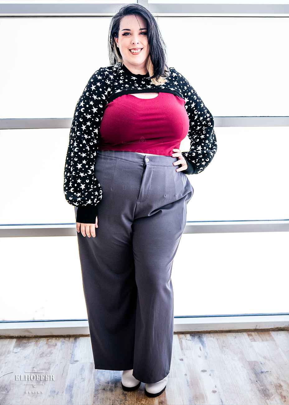 Katie Lynn, a fair skinned 2xl model with black and white hair, is wearing a black super cropped crew neck knit shrug sweater with a white star and moon pattern, long billowing sleeves, and thumbholes