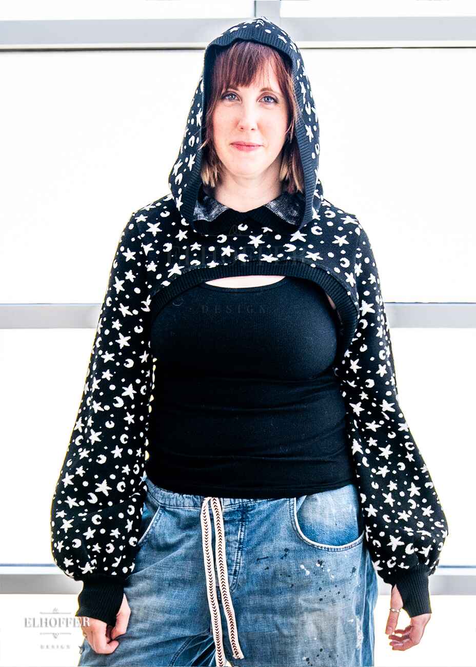 Jes, a fair skinned S model with short red hair and bangs, is wearing the L sample of a black hooded super cropped knit shrug sweater with a white star and moon pattern, long billowing sleeves and thumbholes.