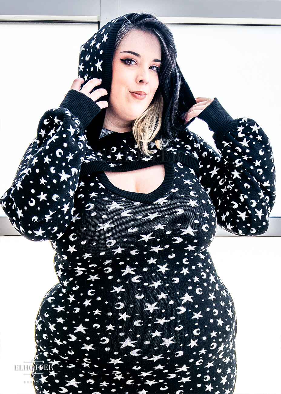 Katie Lynn, a fair skinned 2xl model with black and white hair, is wearing a black hooded super cropped knit shrug sweater with a white star and moon pattern, long billowing sleeves, and thumbholes, paired with a matching fitted knee length bodycon dress.