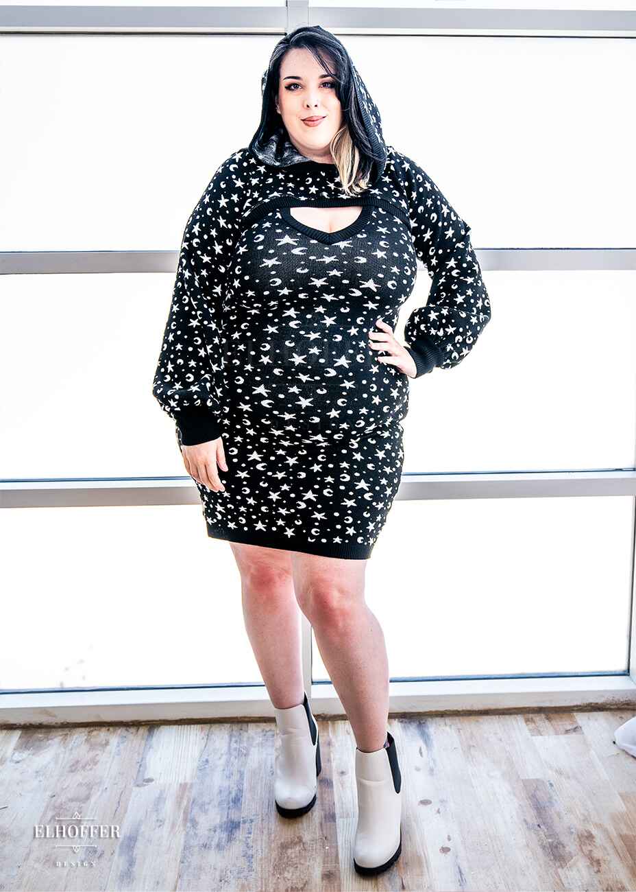 Katie Lynn, a fair skinned 2xl model with black and white hair, is wearing a black hooded super cropped knit shrug sweater with a white star and moon pattern, long billowing sleeves, and thumbholes, paired with a matching fitted knee length bodycon dress.