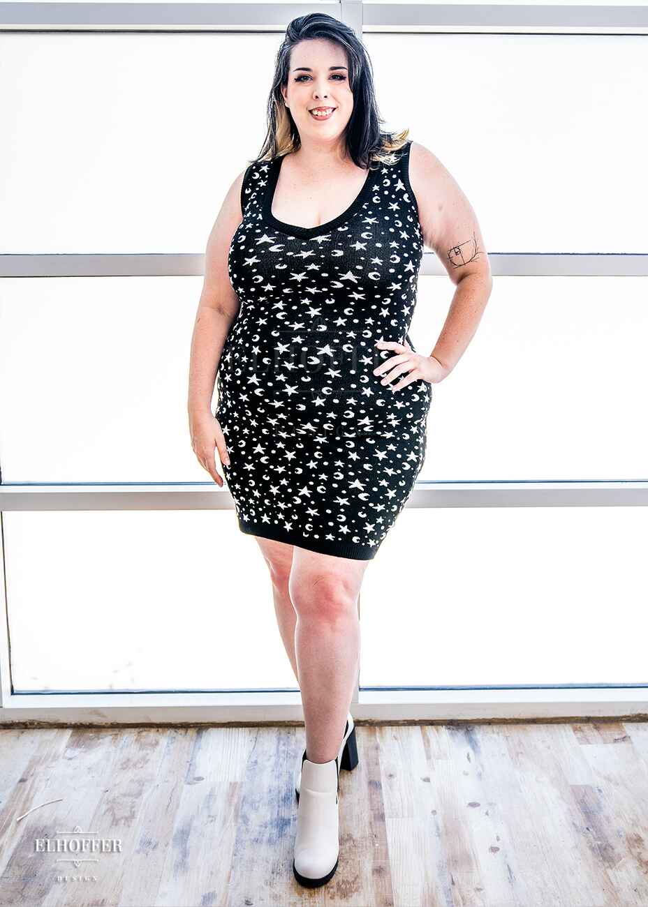 Katie Lynn, a fair skinned 2xl model with black and white hair, is wearing a black fitted knee length knit bodycon dress with a white star and moon pattern and a deep v neckline.