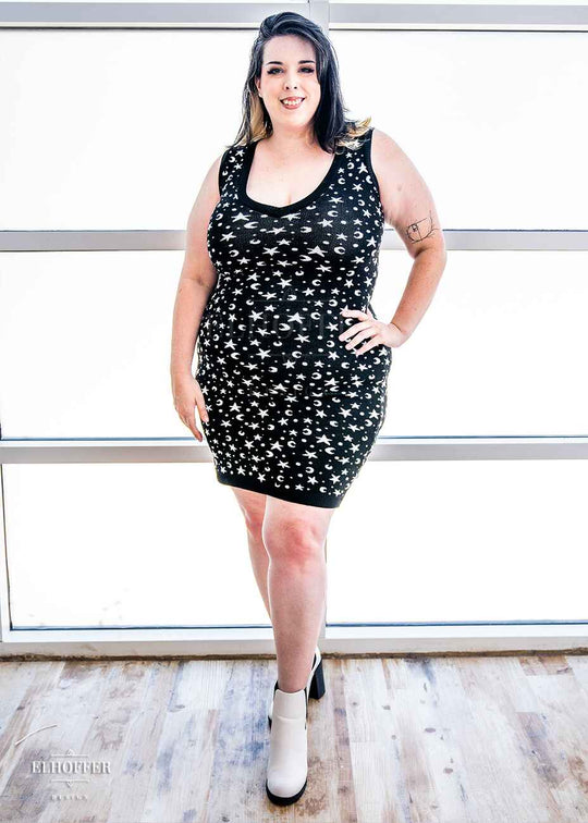 Katie Lynn, a fair skinned 2xl model with black and white hair, is wearing a black fitted knee length knit bodycon dress with a white star and moon pattern and a deep v neckline.