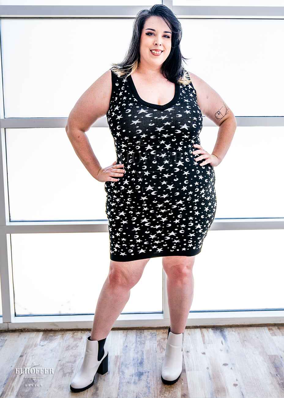 Katie Lynn, a fair skinned 2xl model with black and white hair, is wearing a black fitted knee length knit bodycon dress with a white star and moon pattern and a deep v neckline.