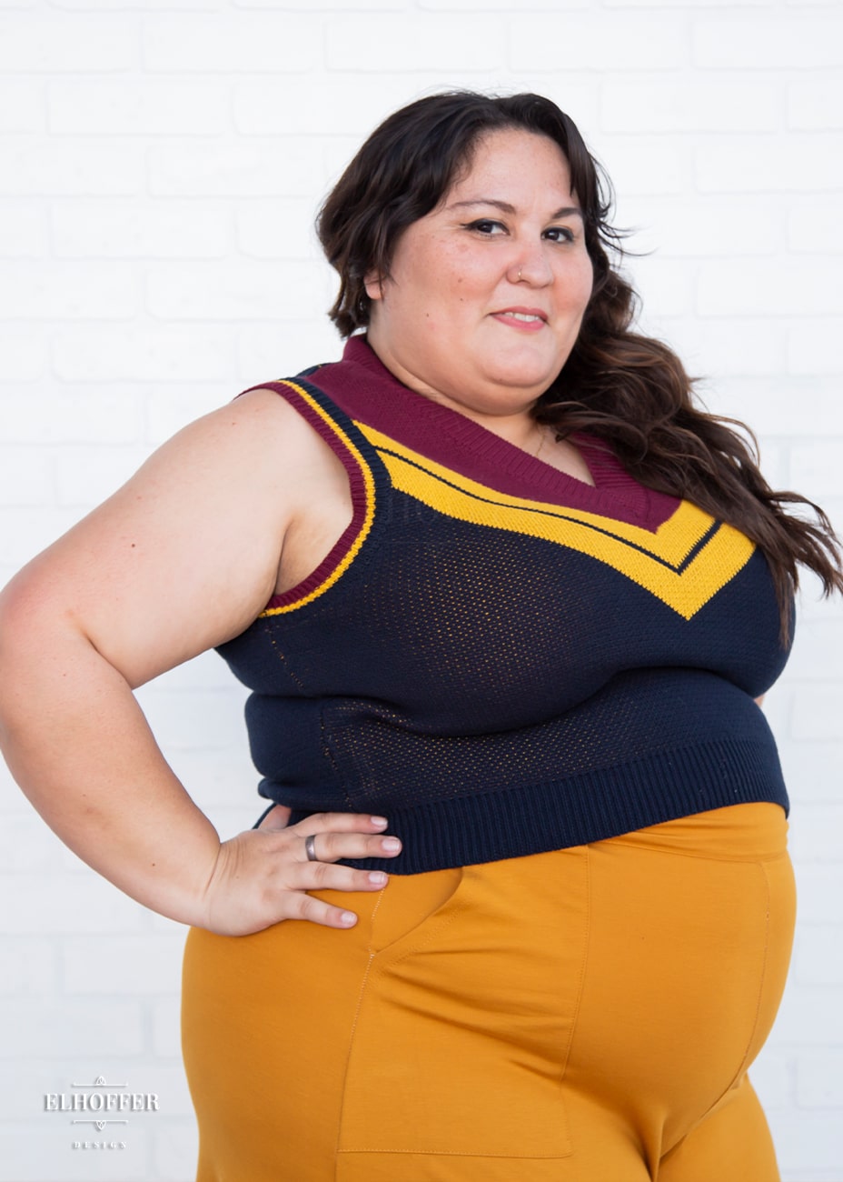 Alysia, a fair skinned size 2XL model with long brown hair, is wearing a pullover vest with a boxy fit and v-neck. It is navy blue with a deep red neckline and mustard yellow v detail.