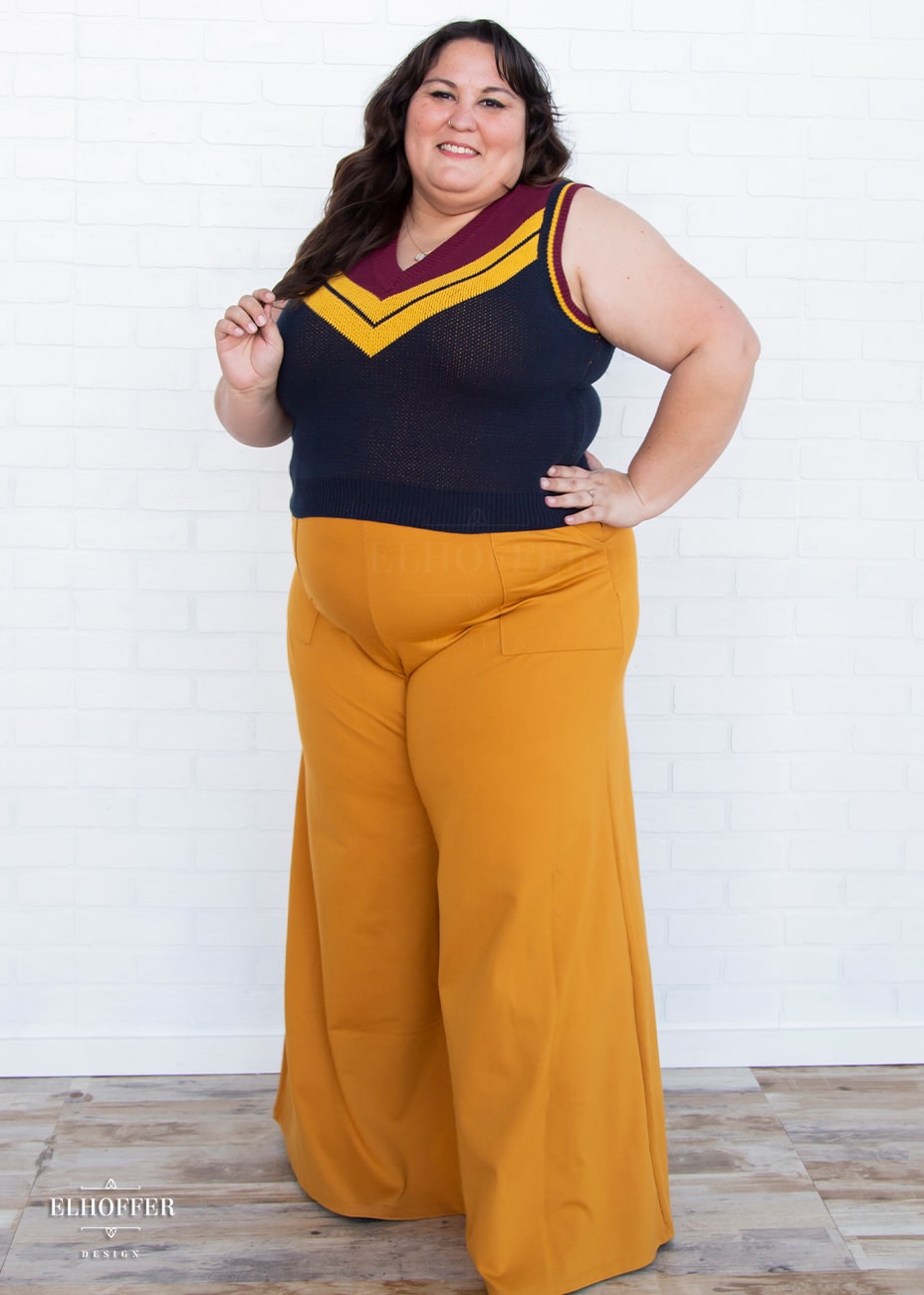 Alysia, a fair skinned size 2XL model with long brown hair, is wearing a pullover vest with a boxy fit and v-neck. It is navy blue with a deep red neckline and mustard yellow v detail.