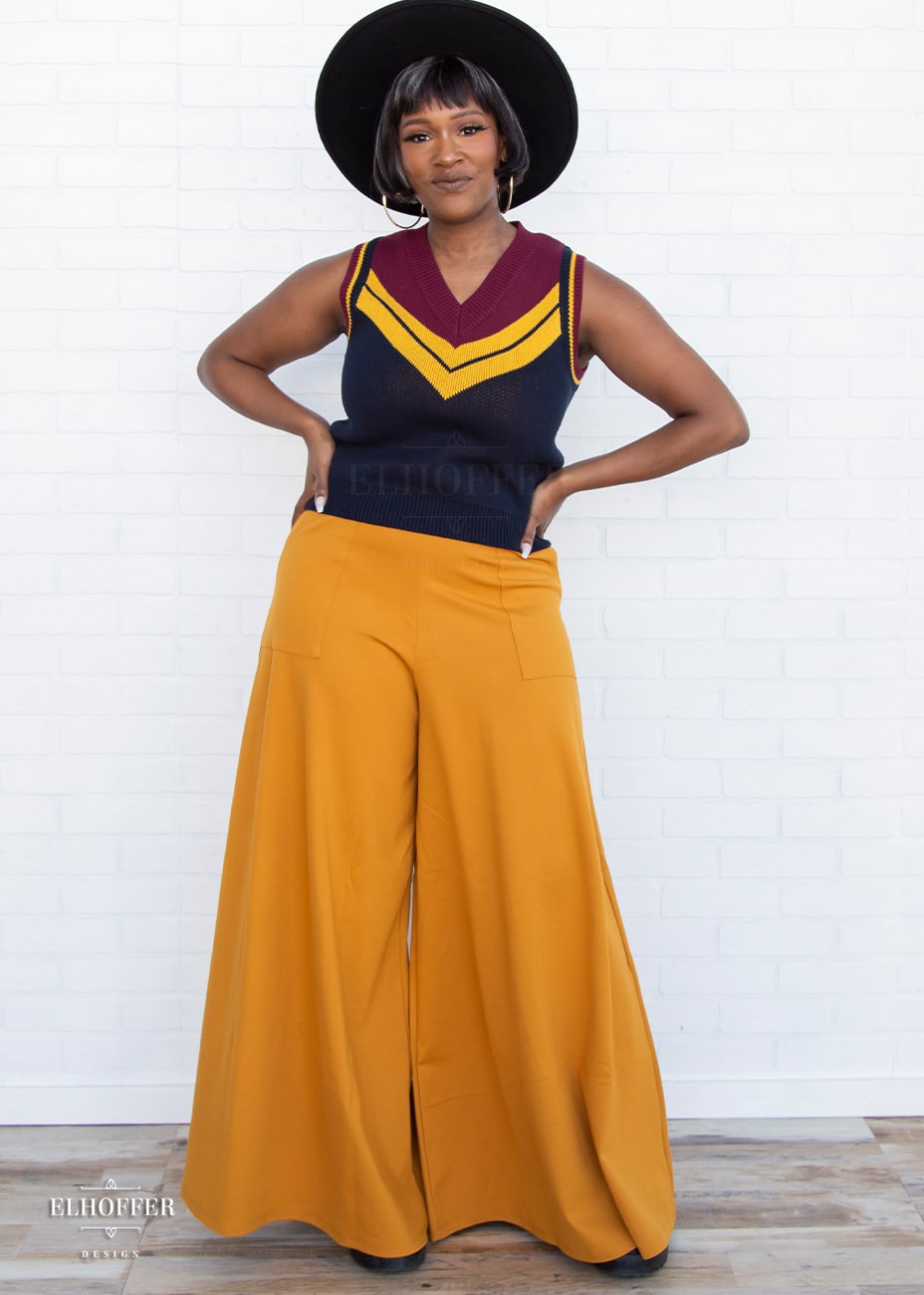 Lynsi, a medium dark skinned model with a dark brown and white bob, is wearing a pullover vest with a boxy fit and v-neck. It is navy blue with a deep red neckline and mustard yellow v detail.