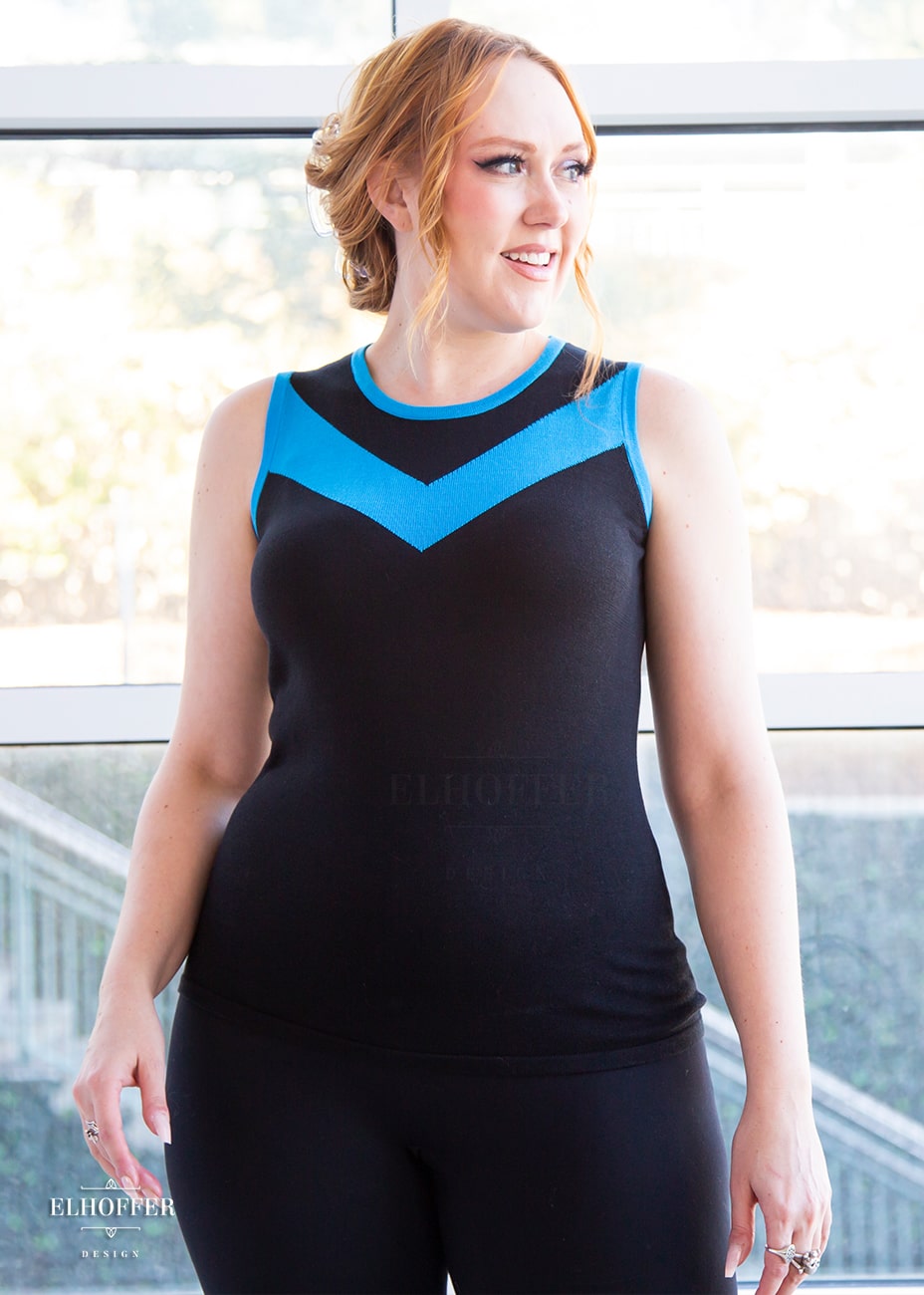 Kelsey, a fair skinned S model with strawberry blonde hair pulled back in a pony tail, is smiling while wearing a light weight sleeveless knit top. The body of the top is mainly black with a bright teal chevron design across the chest and matching teal binding around the neckline and armholes.