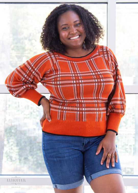 Maydelle, a medium dark skinned XL model with shoulder length curly dark brown hair, is wearing a cropped boatneck sweater with half length bishop sleeve in an orange, white, and black tartan.