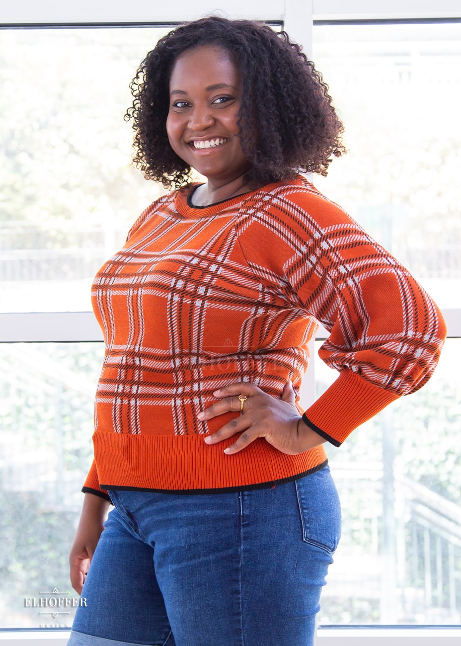 Maydelle, a medium dark skinned XL model with shoulder length curly dark brown hair, is wearing a cropped boatneck sweater with half length bishop sleeve in an orange, white, and black tartan.