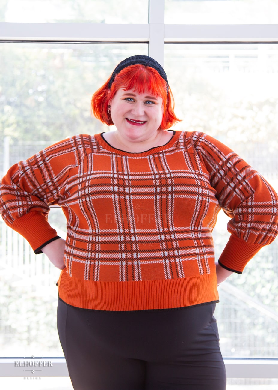 Logan, a fair skinned 3XL model with short bright orange hair and bangs, is wearing a cropped boatneck sweater with half length bishop sleeve in an orange, white, and black tartan.