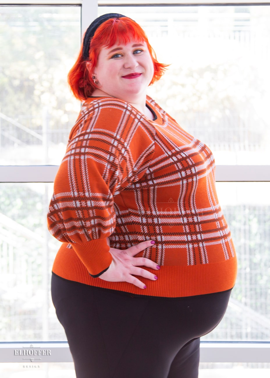 Logan, a fair skinned 3XL model with short bright orange hair and bangs, is wearing a cropped boatneck sweater with half length bishop sleeve in an orange, white, and black tartan.