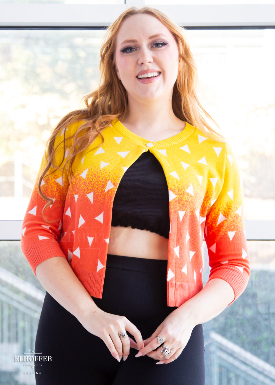Kelsey, a fair skinned size small model with long ginger hair, is wearing a 3/4 sleeved cropped cardigan with one button at the neckline. It is yellow at the top and gradually turns orange toward the bottom. It has white triangles throughout.