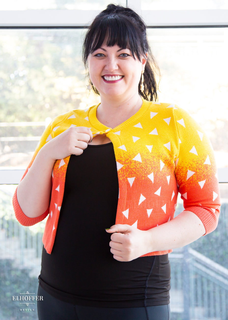 Bernadette, a fair skinned size large model with dark hair and bangs, is wearing a 3/4 sleeved cropped cardigan with one button at the neckline. It is yellow at the top and gradually turns orange toward the bottom. It has white triangles throughout.