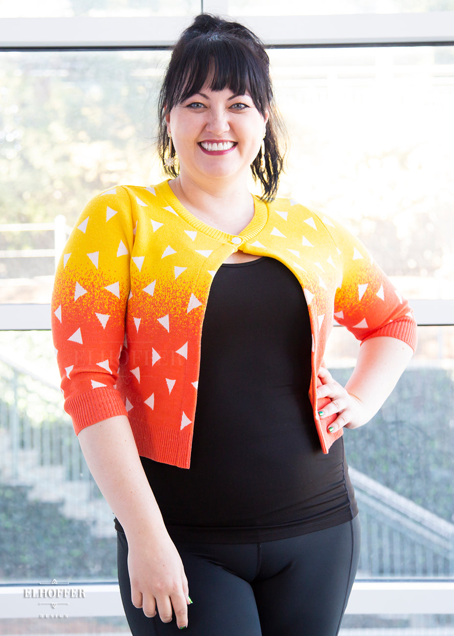 Bernadette, a fair skinned size large model with dark hair and bangs, is wearing a 3/4 sleeved cropped cardigan with one button at the neckline. It is yellow at the top and gradually turns orange toward the bottom. It has white triangles throughout.