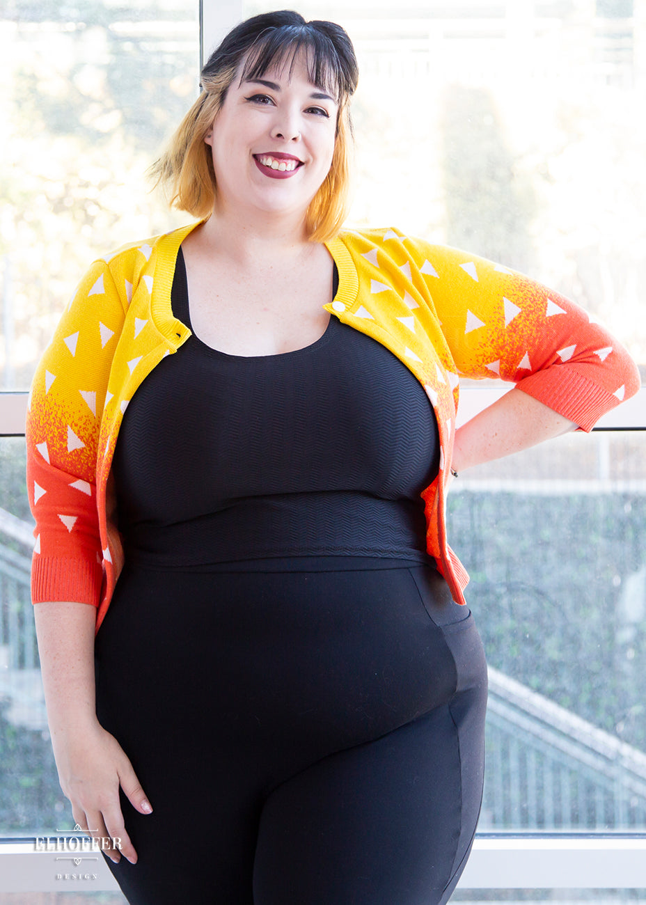Katie Lynn, a fair skinned size 2XL model with short black and blonde hair, is wearing a 3/4 sleeved cropped cardigan with one button at the neckline. It is yellow at the top and gradually turns orange toward the bottom. It has white triangles throughout.