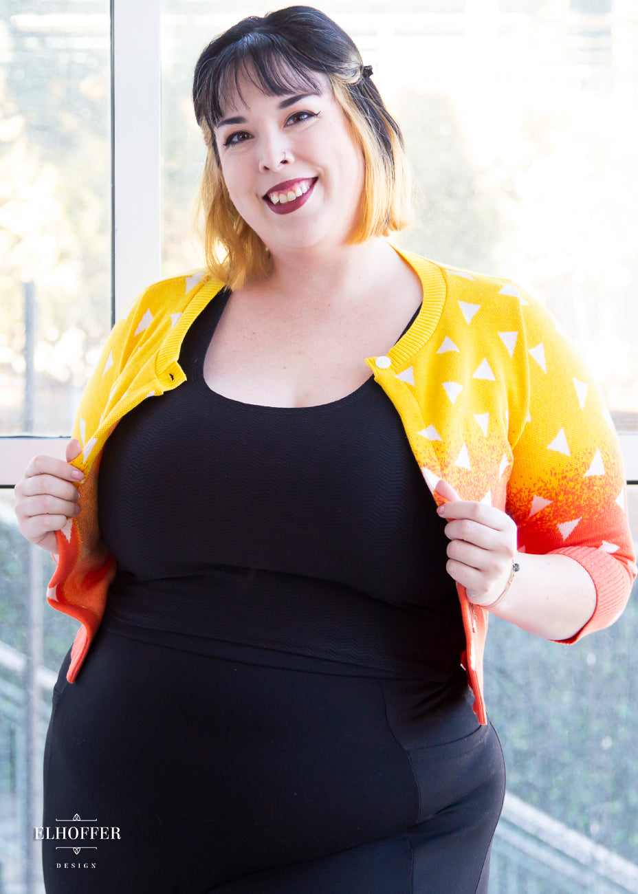 Katie Lynn, a fair skinned size 2XL model with short black and blonde hair, is wearing a 3/4 sleeved cropped cardigan with one button at the neckline. It is yellow at the top and gradually turns orange toward the bottom. It has white triangles throughout.