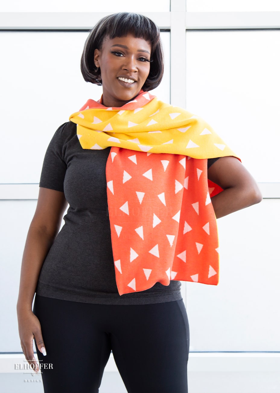 Lynsi, a medium dark skinned M model with short black and white hair, is smiling while wearing a reversible knit scarf.  One side of the scarf is orange with white triangles and the other side is yellow with white triangles.