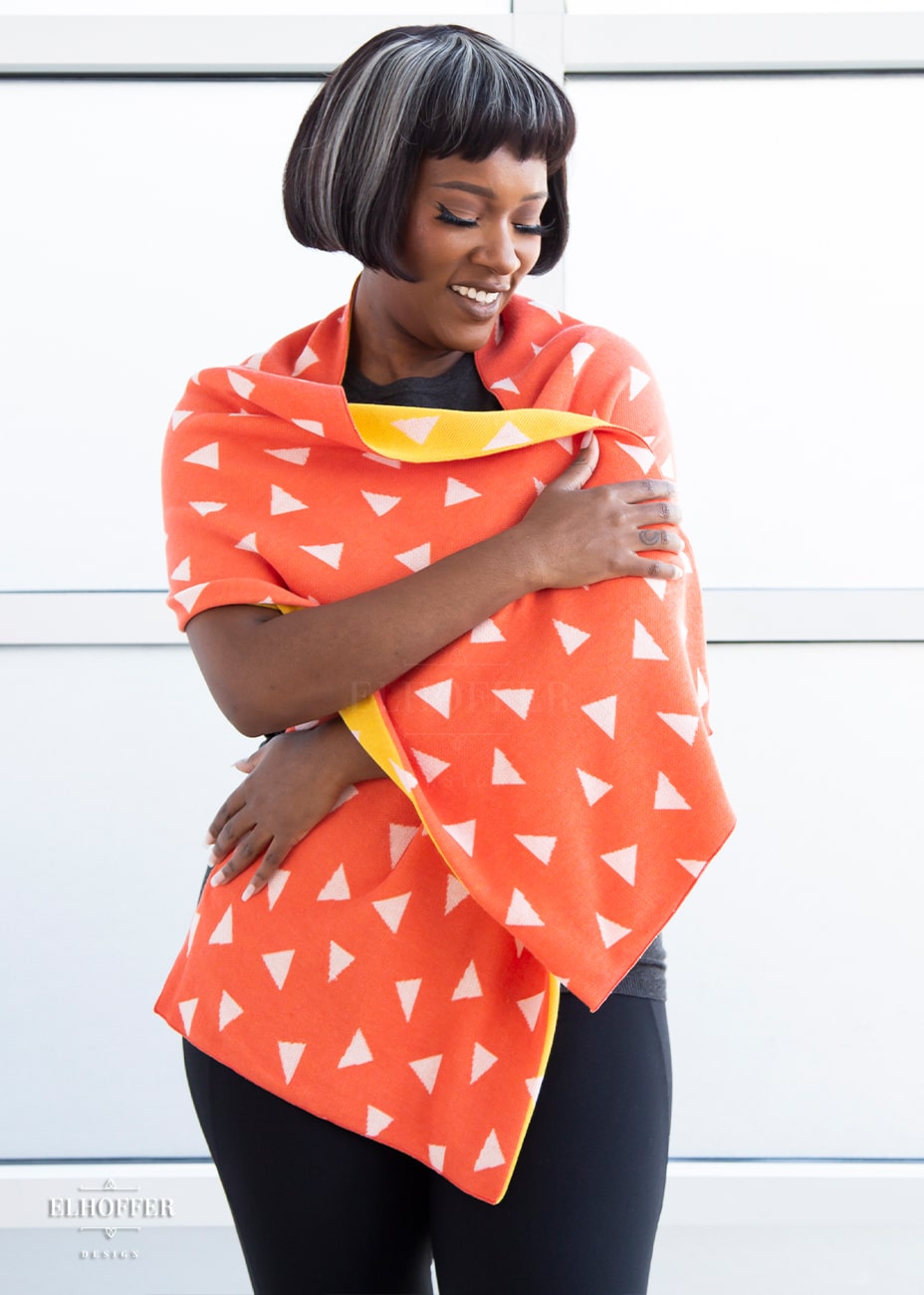 Lynsi, a medium dark skinned M model with short black and white hair, is smiling while wrapped up in a reversible knit scarf.  One side of the scarf is orange with white triangles and the other side is yellow with white triangles.