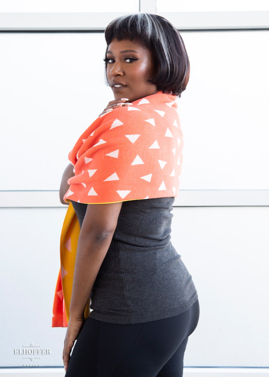 Lynsi, a medium dark skinned M model with short black and white hair, is wearing a reversible knit scarf.  One side of the scarf is orange with white triangles and the other side is yellow with white triangles.
