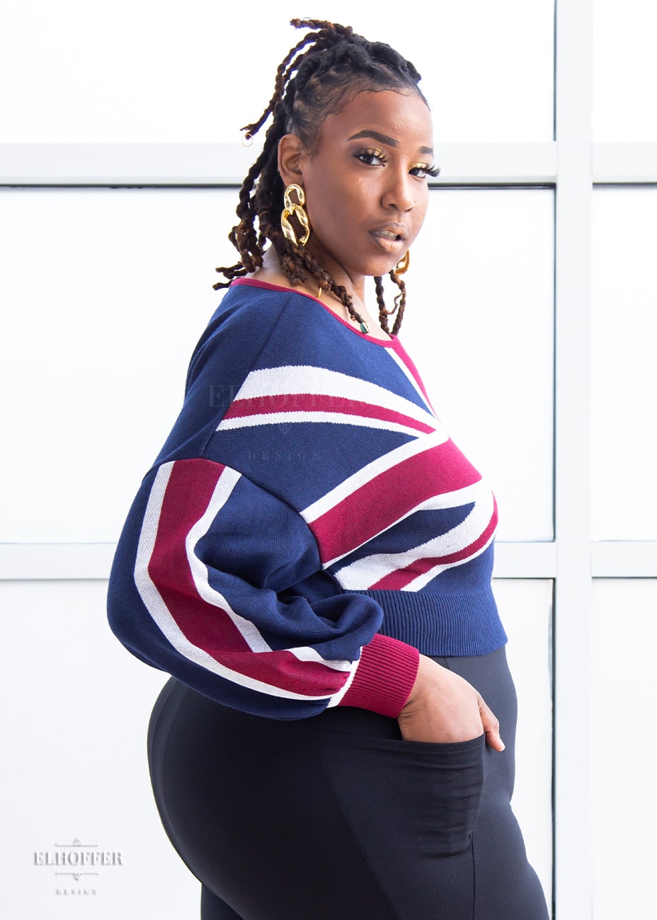 Myjah, a medium dark skinned 3xl model with braids, is wearing a cropped oversize sweater with a Union Jack design and billowing sleeves paired with black leggings. 