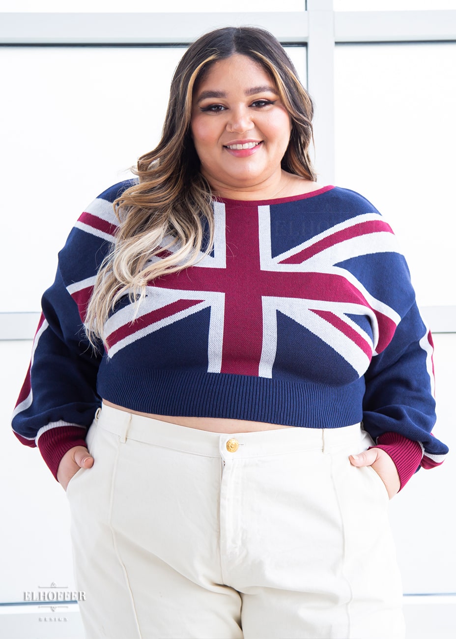 Cori, a sun kissed skinned 2xl model with long balayage hair, is smiling while wearing a cropped oversize sweater with a Union Jack design and long billowing sleeves.